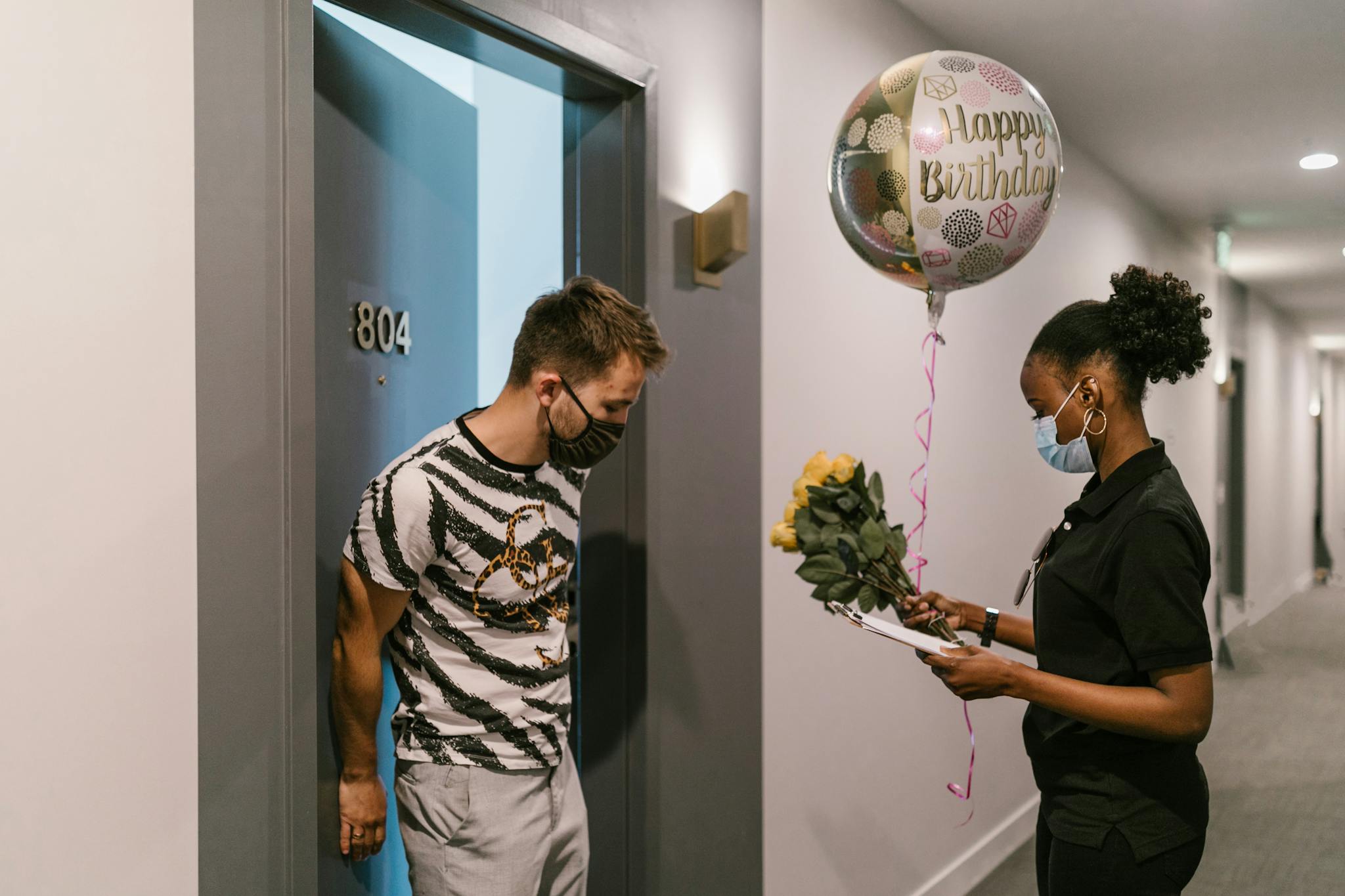A deliverywoman brings a birthday surprise with flowers and a balloon to a masked recipient at their apartment.