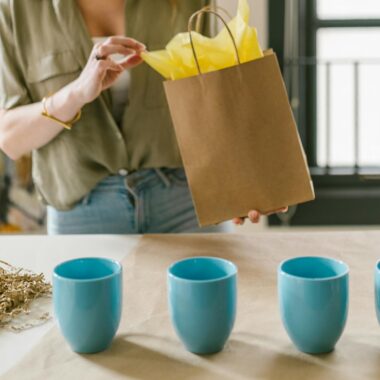 A woman packing a brown paper bag with colorful tissue alongside blue cups on a table.