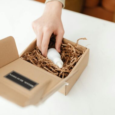 Close-up of a hand unpacking a cardboard box with a product bottle inside on a white table.